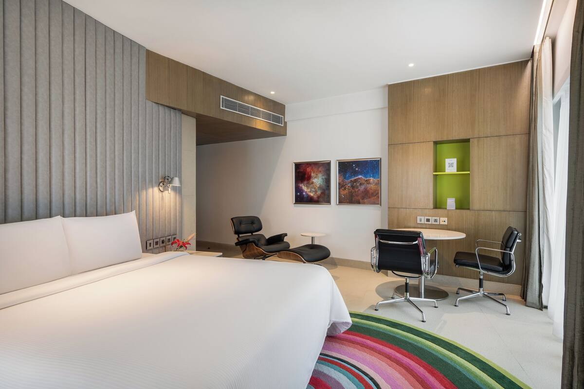 A modern hotel room with a large bed, a desk, two chairs, and a colorful rug.