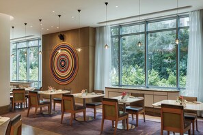 A modern dining area with multiple tables and chairs, large windows with a view of greenery, and a colorful circular art piece on the wall. - Avasa Hotel (Hyderabad)