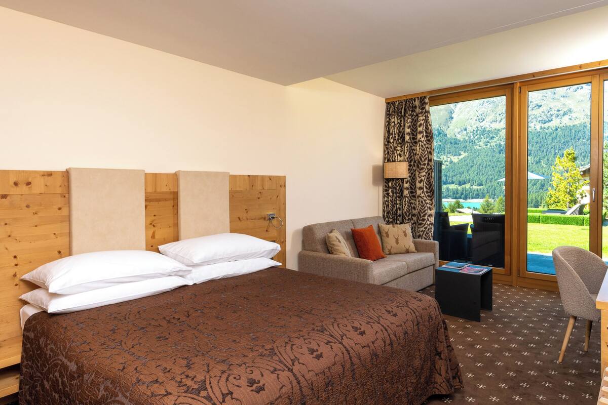 Een hotelkamer met een groot bed, een bank, een klein tafeltje en uitzicht op een bergachtig landschap door een groot raam.