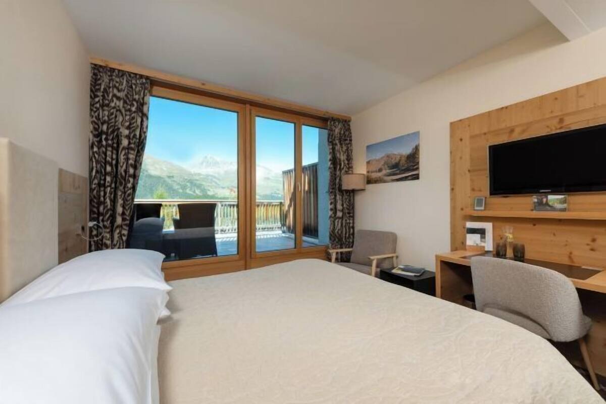 Een hotelkamer met een groot bed, een bureau met stoel, een televisie en een balkon met gordijnen.