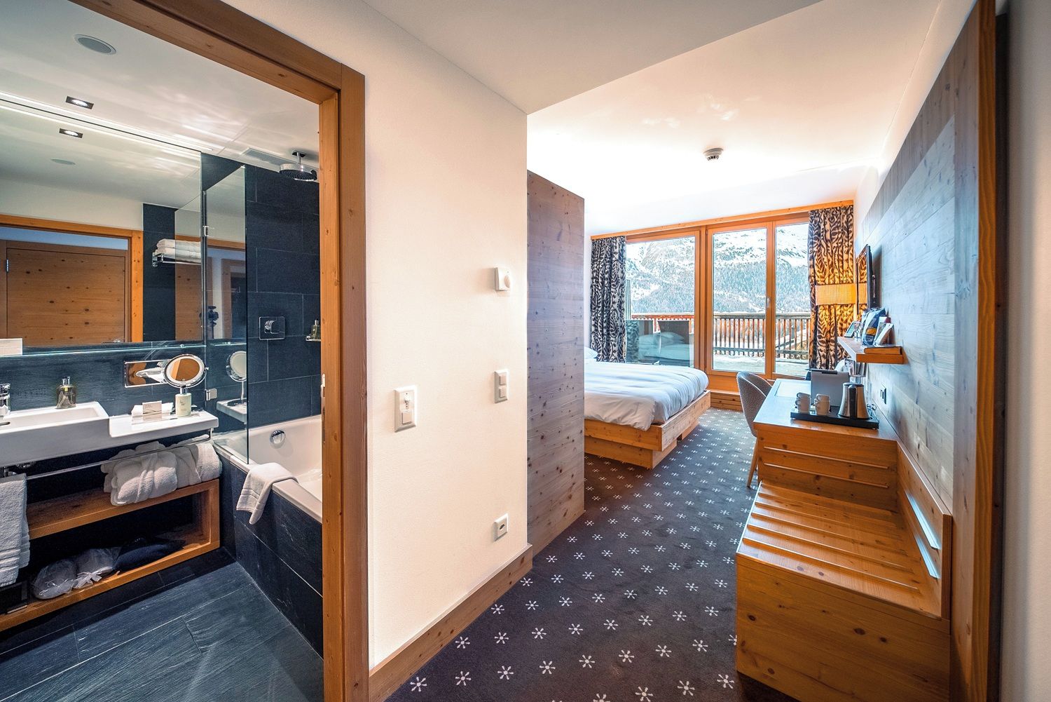 A modern bedroom with a large window, a wooden staircase, a bathroom with a mirror and sink, and a bed with a view of the outdoors.