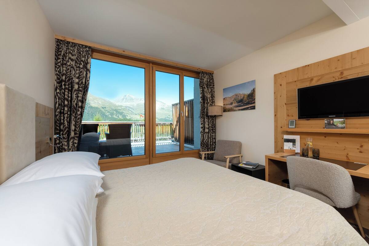 Hotelkamer met groot bed, bureau, stoel, televisie en balkon met uitzicht op de bergen.