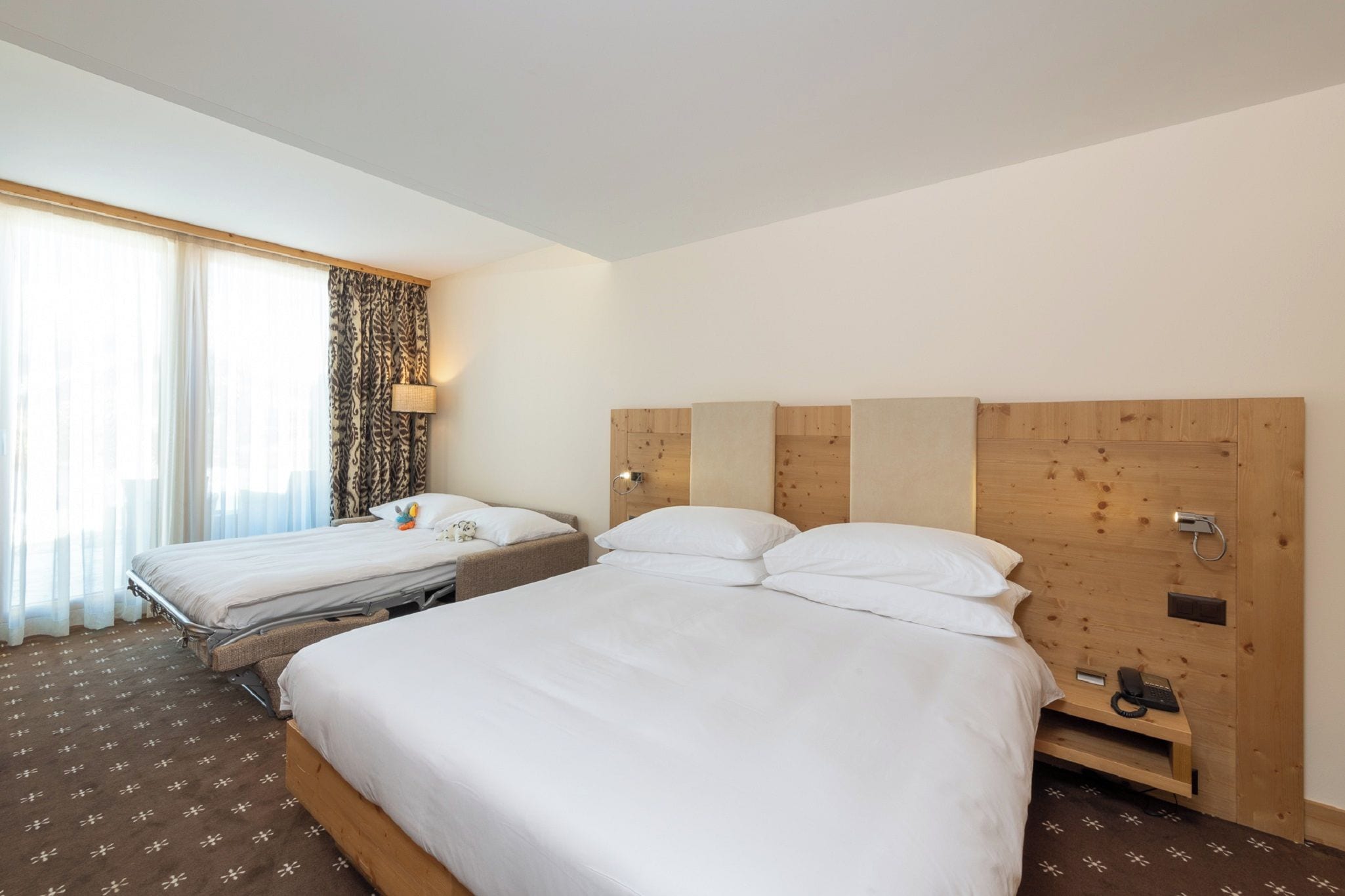 Habitación de hotel con dos camas, una ventana con cortinas traslúcidas, una cortina estampada, una lámpara de noche y un cabecero de madera.