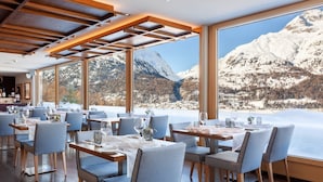 Un restaurant avec des tables dressées pour le service, de grandes fenêtres offrant une vue sur les montagnes et une structure de plafond en bois.