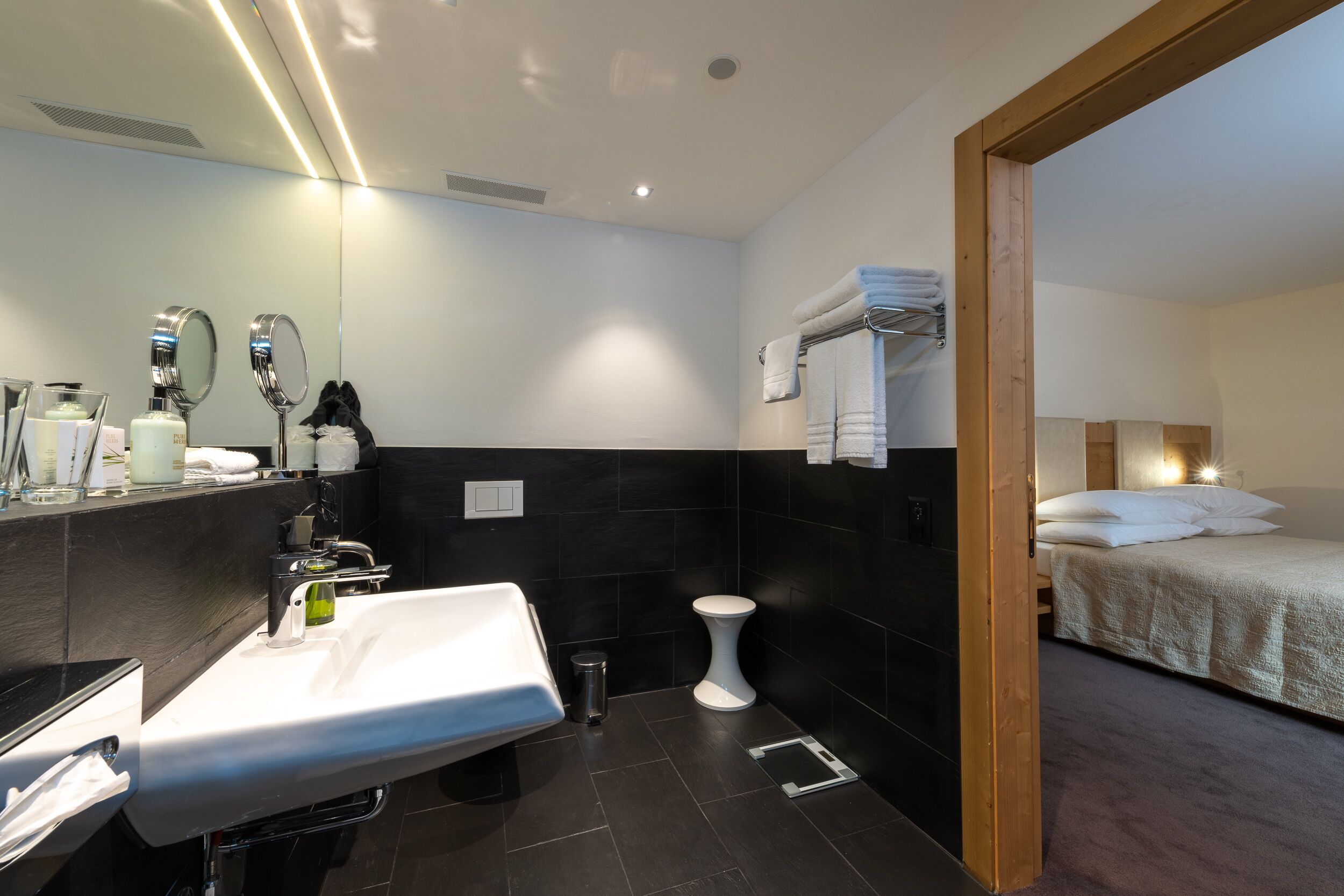 A modern bathroom with a sink, mirrors, and a toilet. There is a bed with white bedding in the adjacent room.