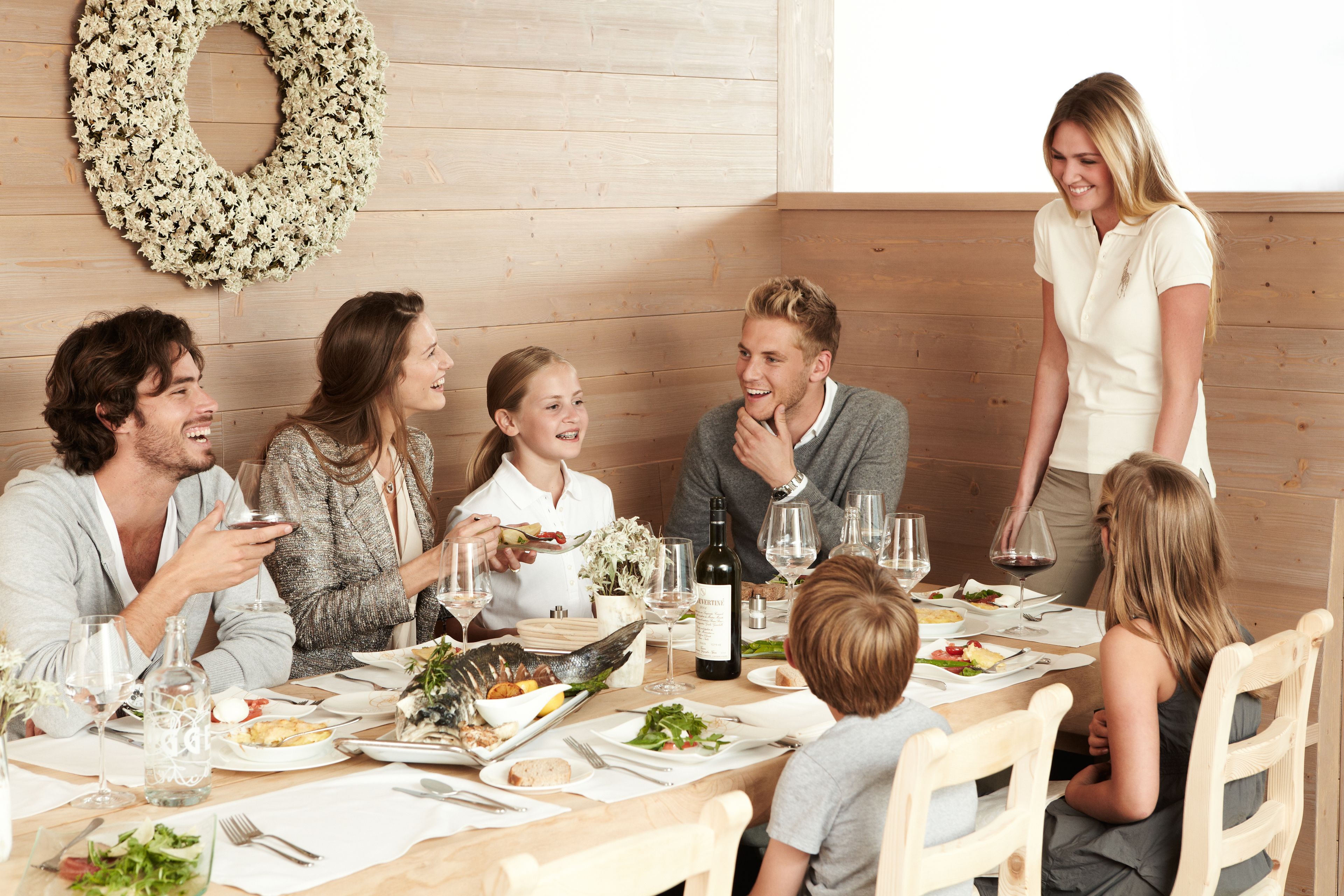 A family dining at a wooden table with a wreath on the wall, surrounded by chairs and a wooden bench.