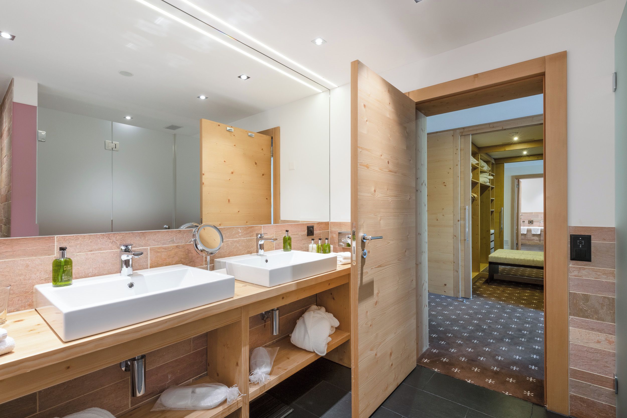 A modern bathroom with two sinks, a mirror, and a wooden door leading to another room.