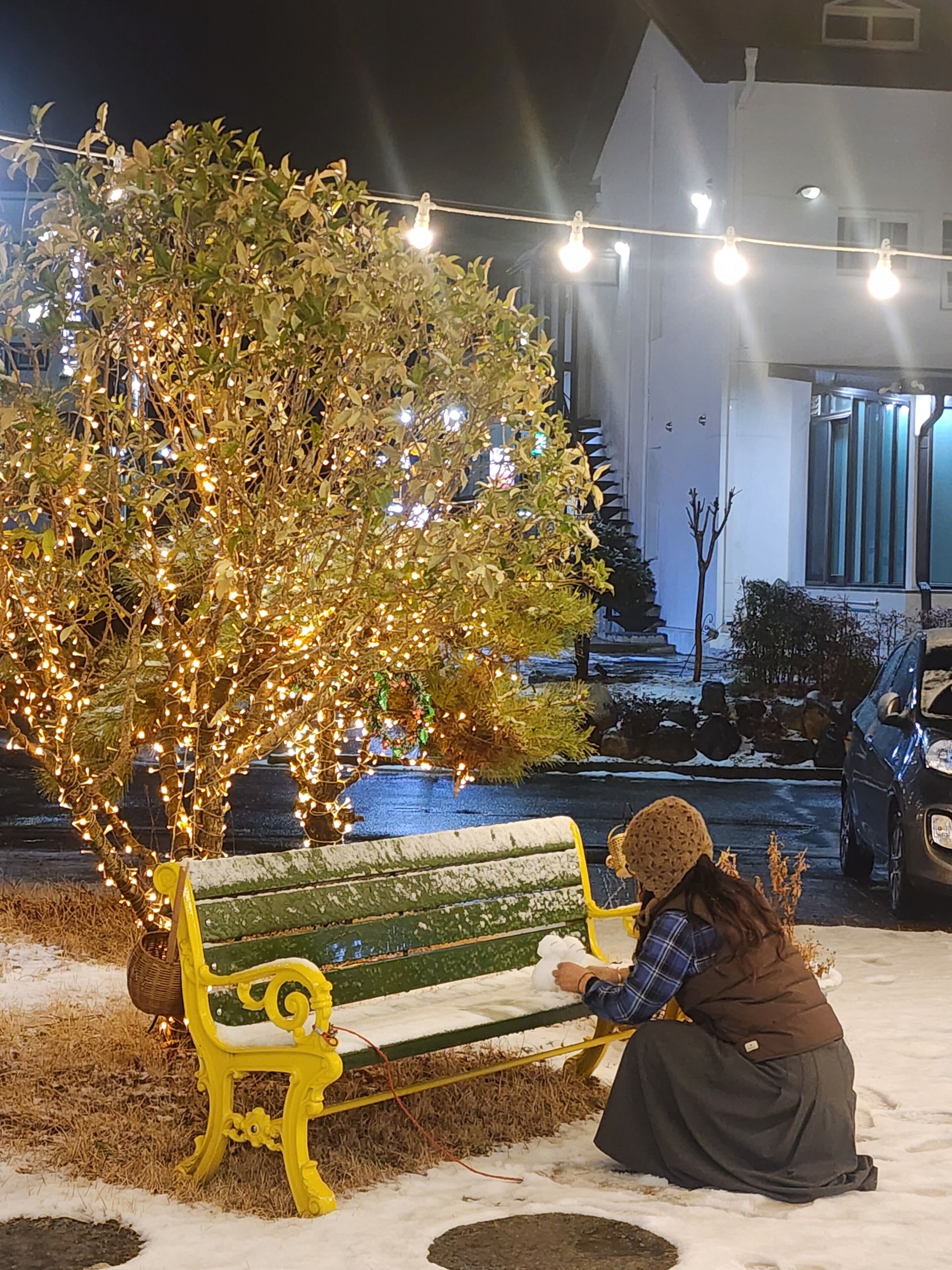 Eine Bank mit einer sitzenden Person, ein Baum mit Lichtern und ein Gebäude im Hintergrund.