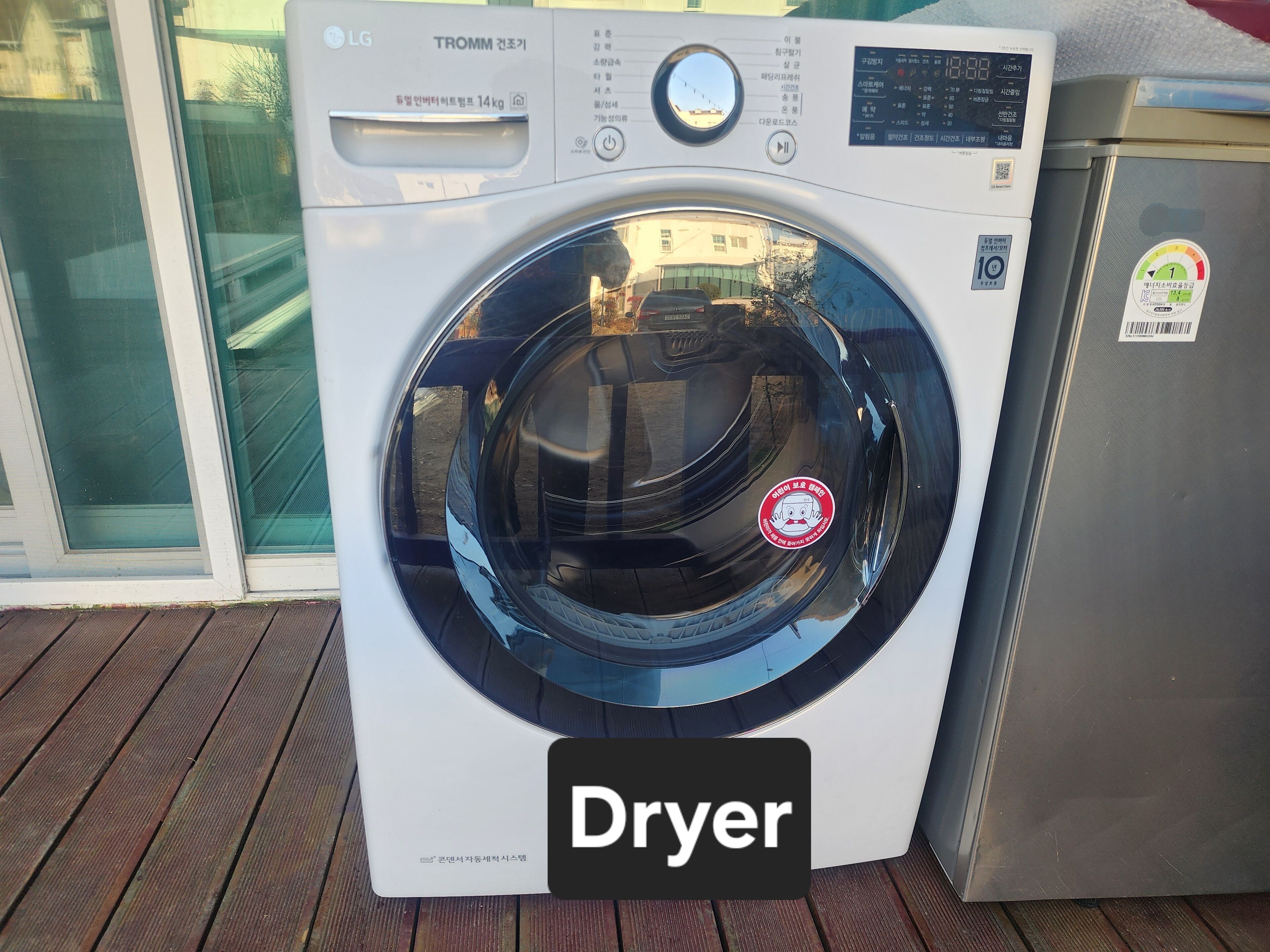A front view of a white LG TROMM dryer with a digital display and control panel, placed on a wooden deck.