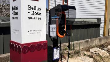 Una estación de carga pública con el rótulo 'Bellus Rose', un número de contacto y un edificio cercano.