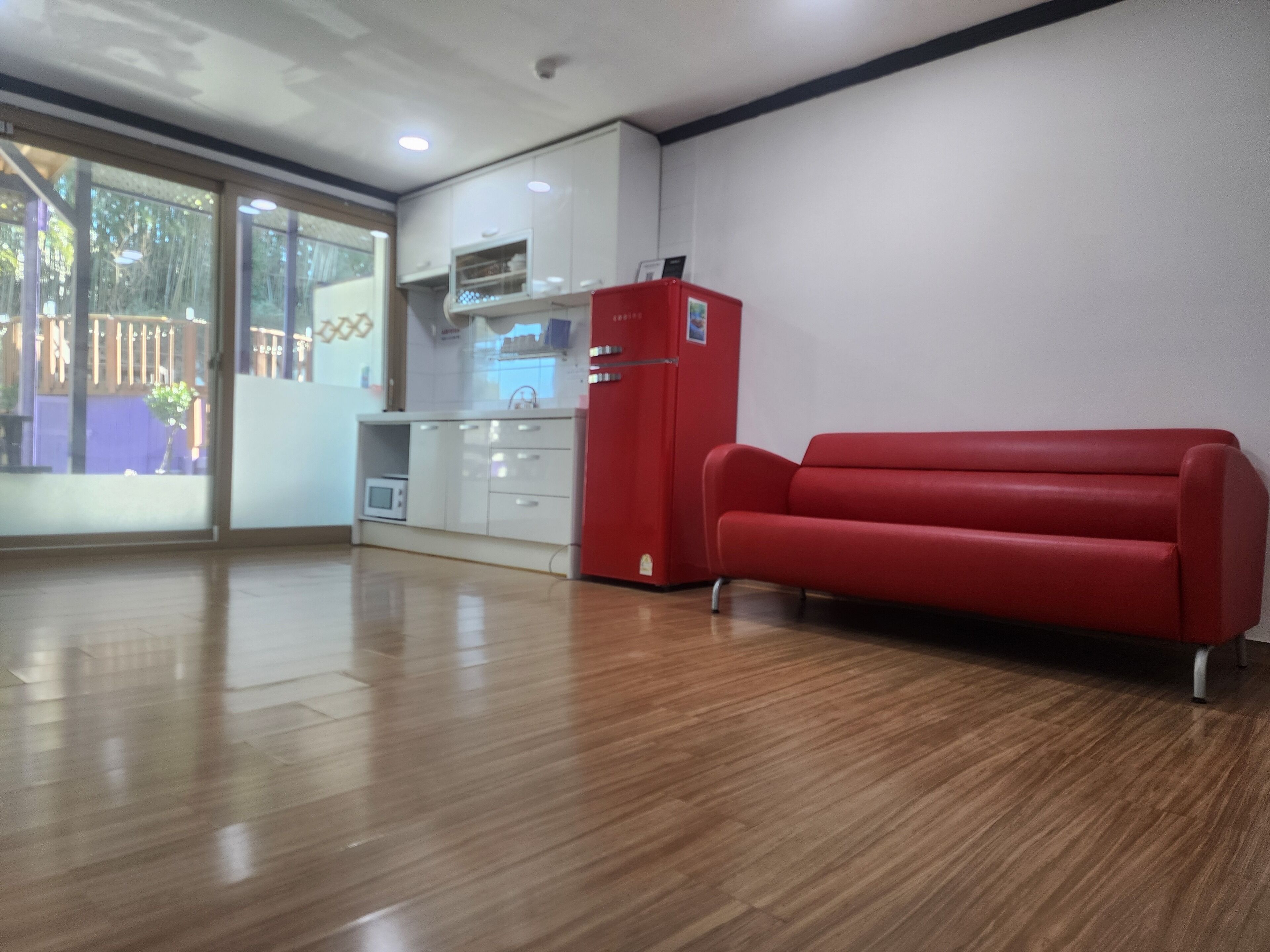 A spacious room with a wooden floor, a red sofa, a white kitchenette, and a sliding glass door leading to an outdoor area.