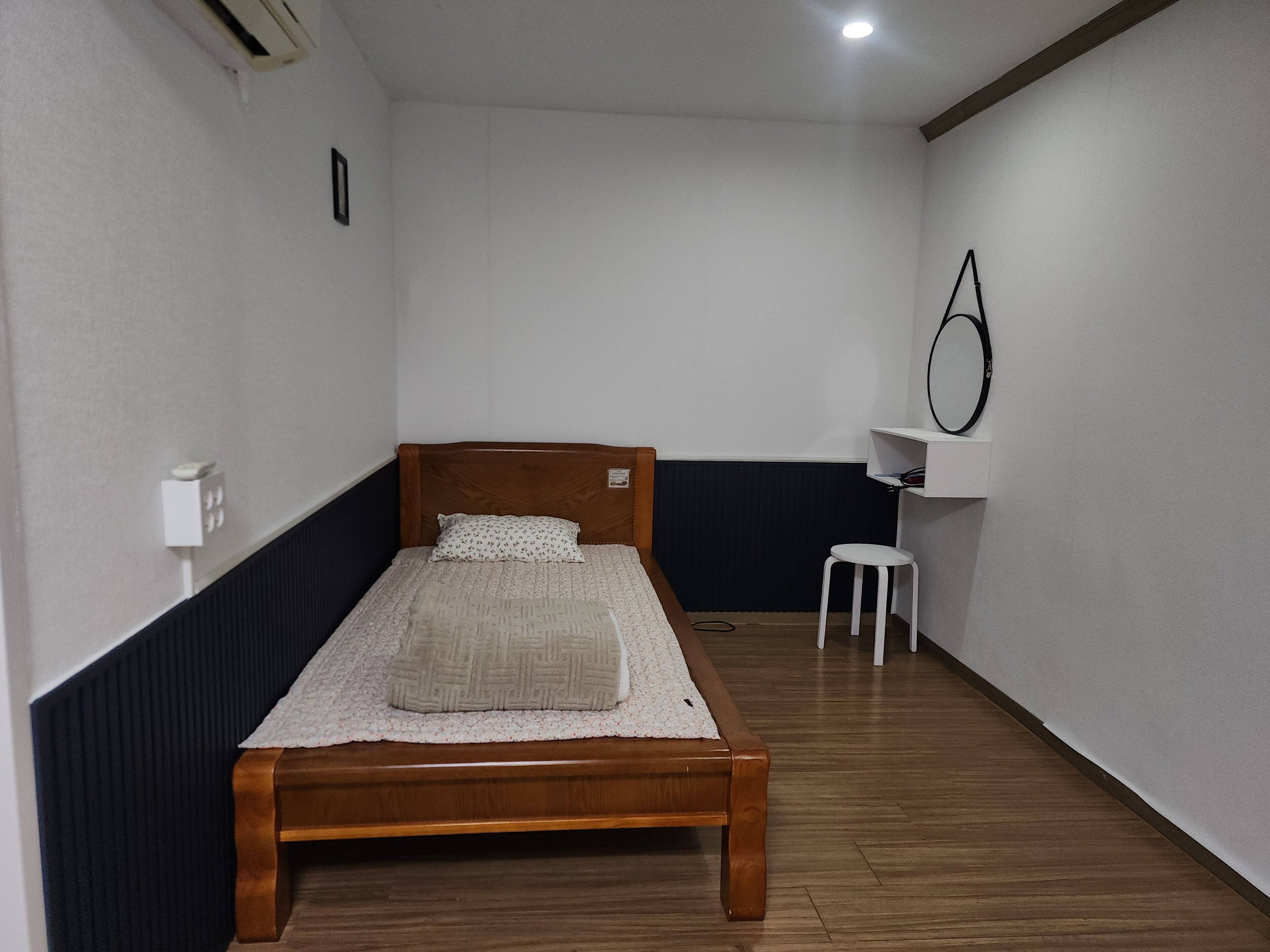 A single bed with a wooden frame and a white mattress, a small white stool, a wall-mounted mirror, and a white shelf with a black circular object.