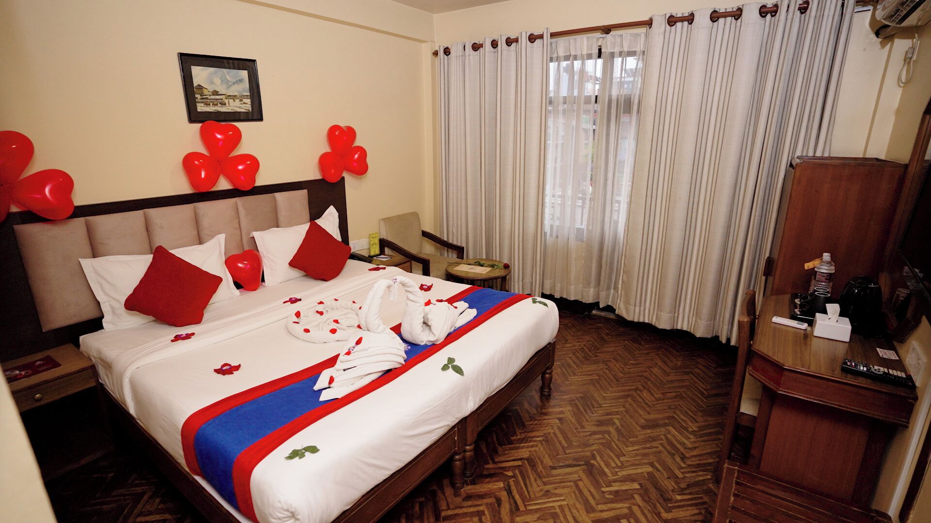 Una habitación de hotel con cama, cabecero de madera, mesita de noche de madera, cómoda de madera, una silla, una pequeña mesa y una ventana con cortinas.