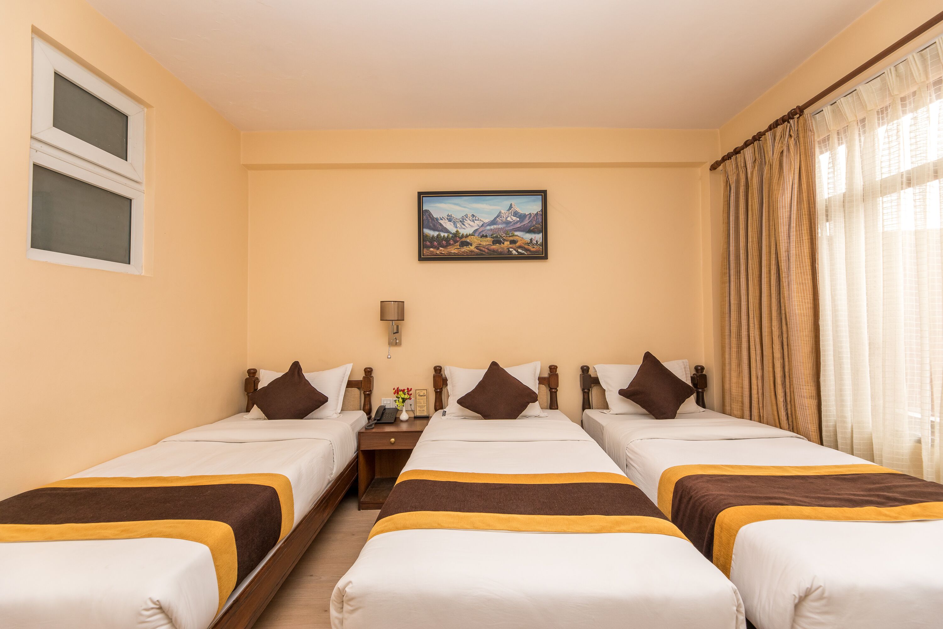 Een hotelkamer met drie enkele bedden, een nachtkastje met een lamp, een schilderij aan de muur en een raam met gordijnen.