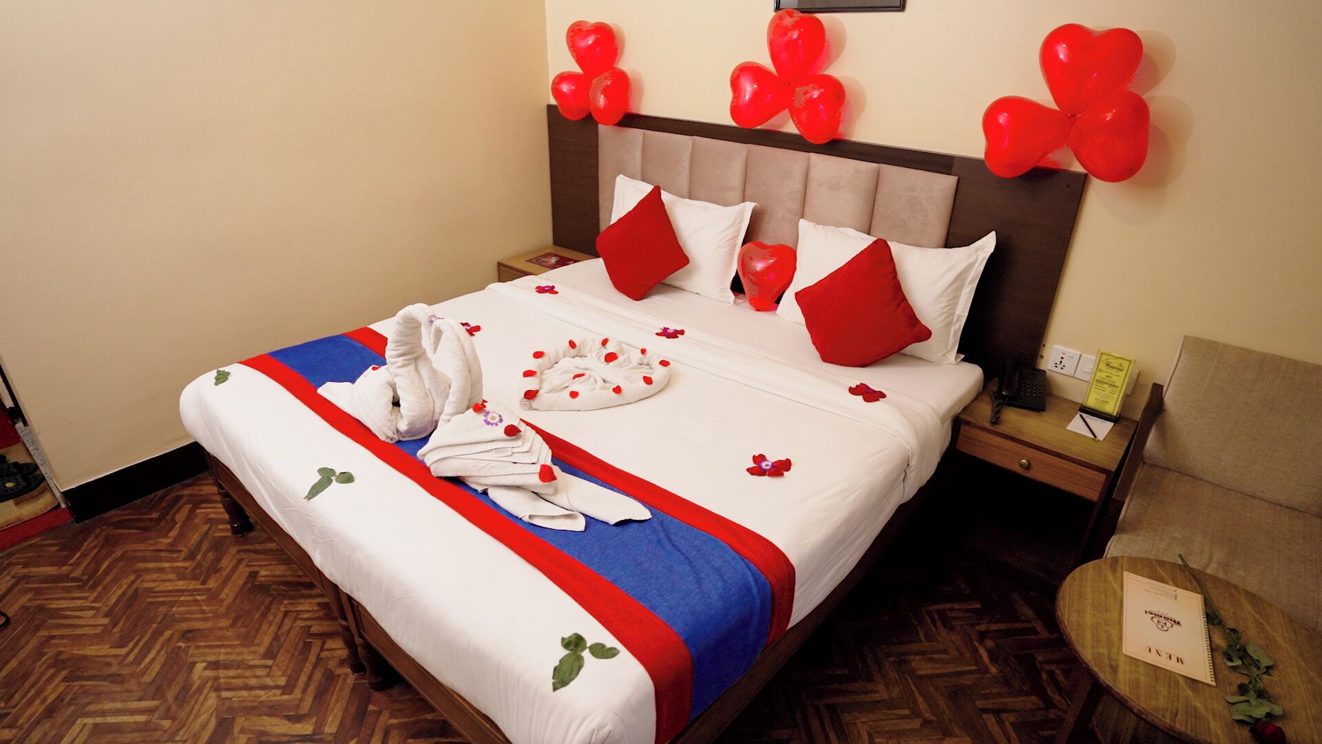Ein ordentlich gemachteres Bett mit einer rot-weiß gestreiften Tagesdecke, herzförmigen roten Kissen und einem Rosenstrauß auf dem Bett.