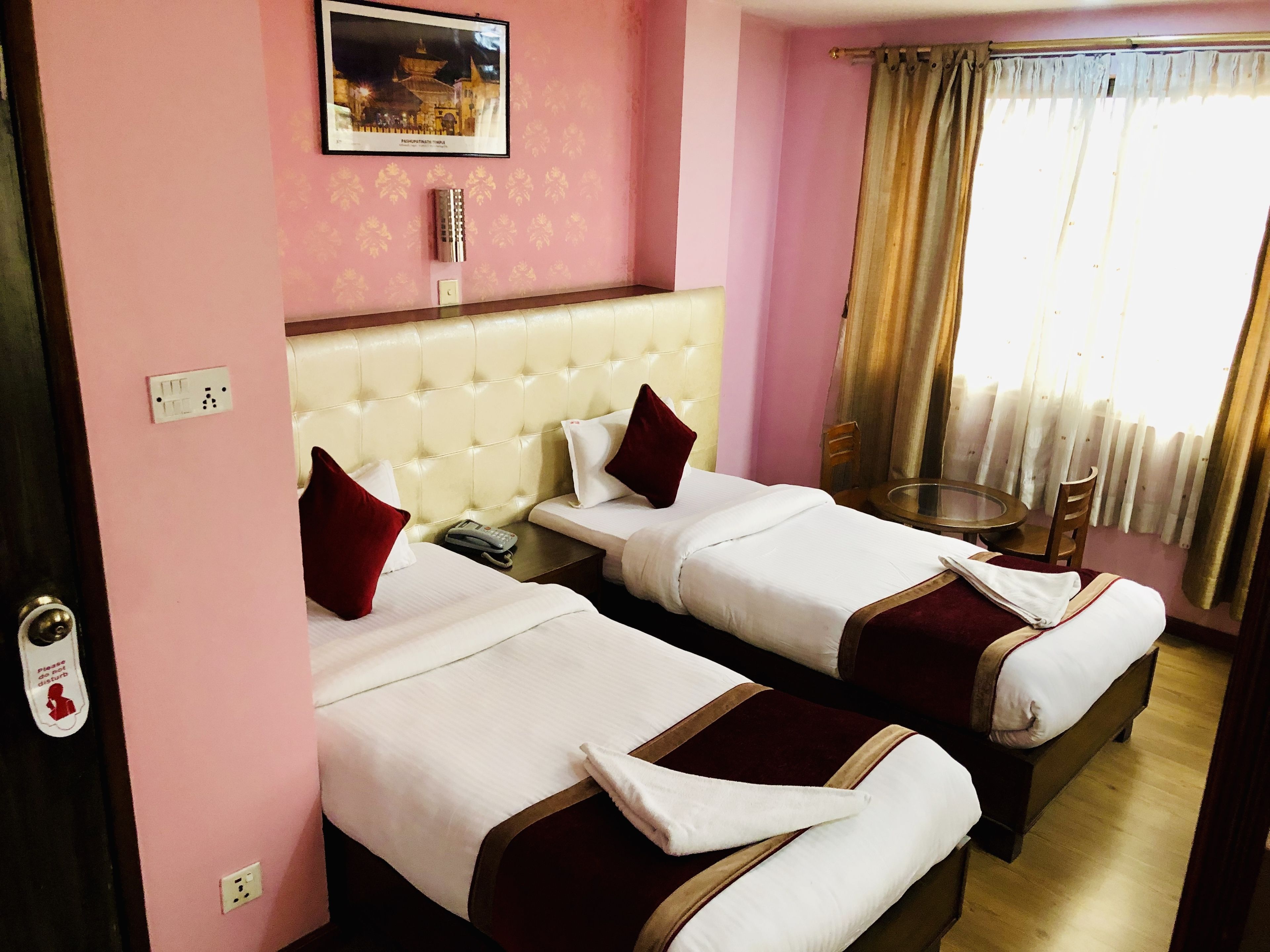Ein Hotelzimmer mit zwei Betten, einem kleinen Tisch, einem Sessel, einem Bild an der Wand und einem Fenster mit Vorhängen.
