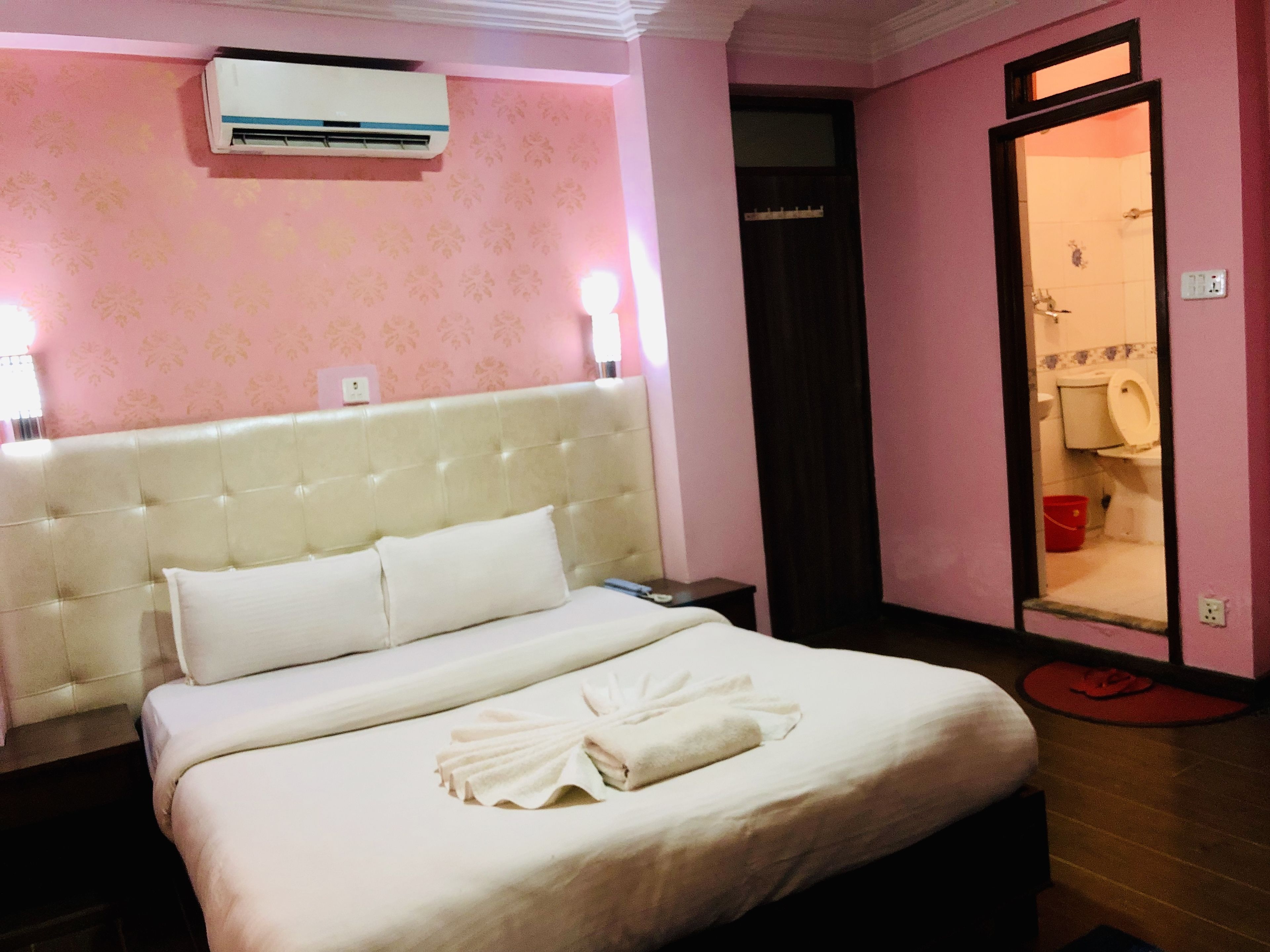 A hotel room with a large bed, a bedside table, an air conditioning unit, and a bathroom visible through an open door.