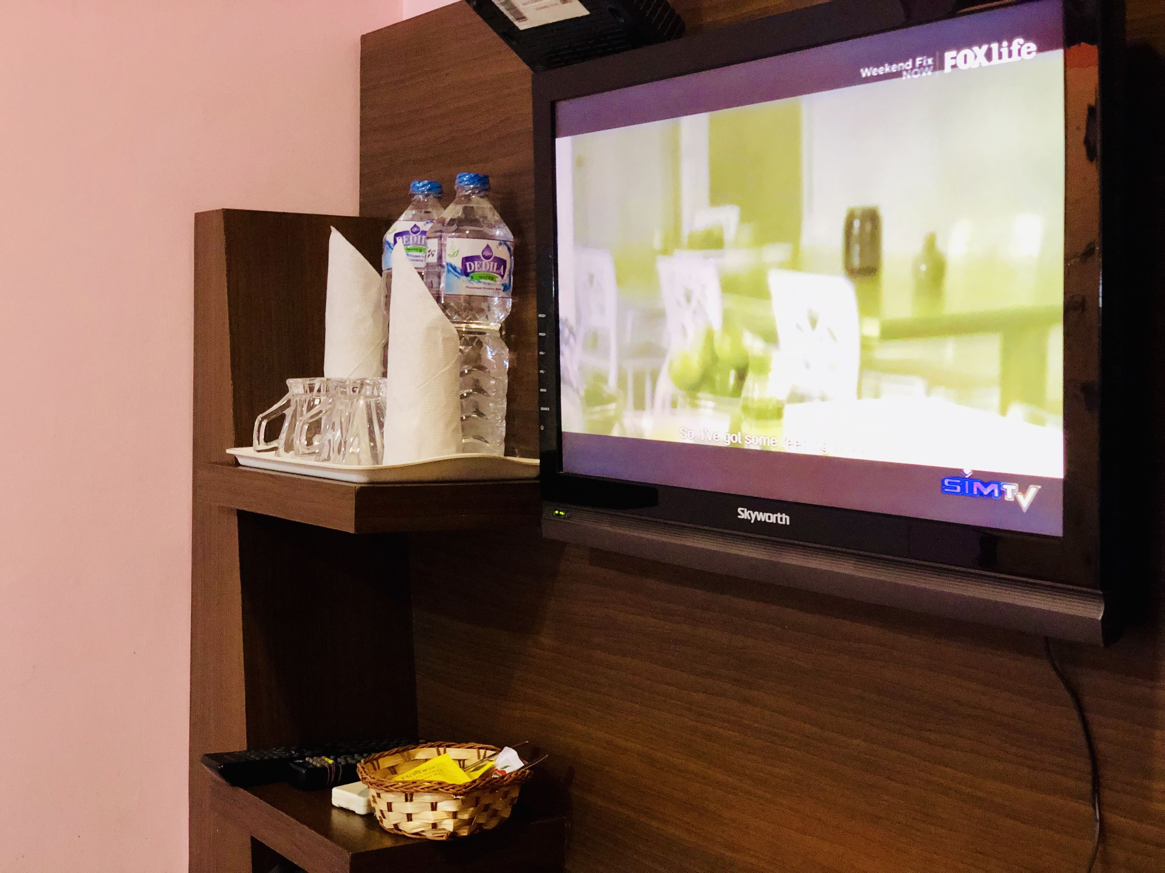 A television displaying a program, a shelf with bottled water, glasses, and a basket with a napkin.