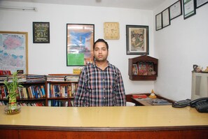 Um homem está de pé em frente a uma escrivaninha de madeira numa sala com estante, quadros e diplomas na parede.