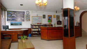 Un área de recepción con un escritorio de madera, una estantería, una zona de estar con sofá y sillones, y varios carteles informativos en las paredes.
