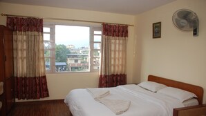 Habitación de hotel con una cama grande, dos ventanas con cortinas, suelo de madera, una mesita de noche, una silla y un ventilador de pared.