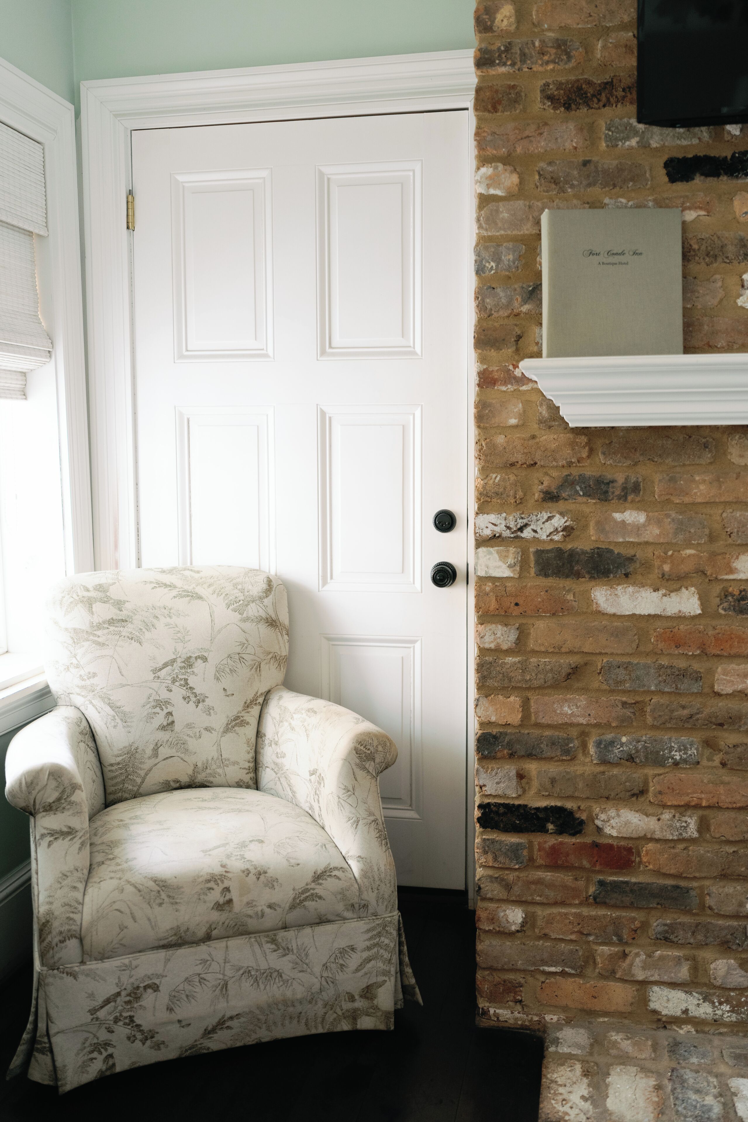 A room with a white door, a floral armchair, a brick wall, and a book on a shelf.