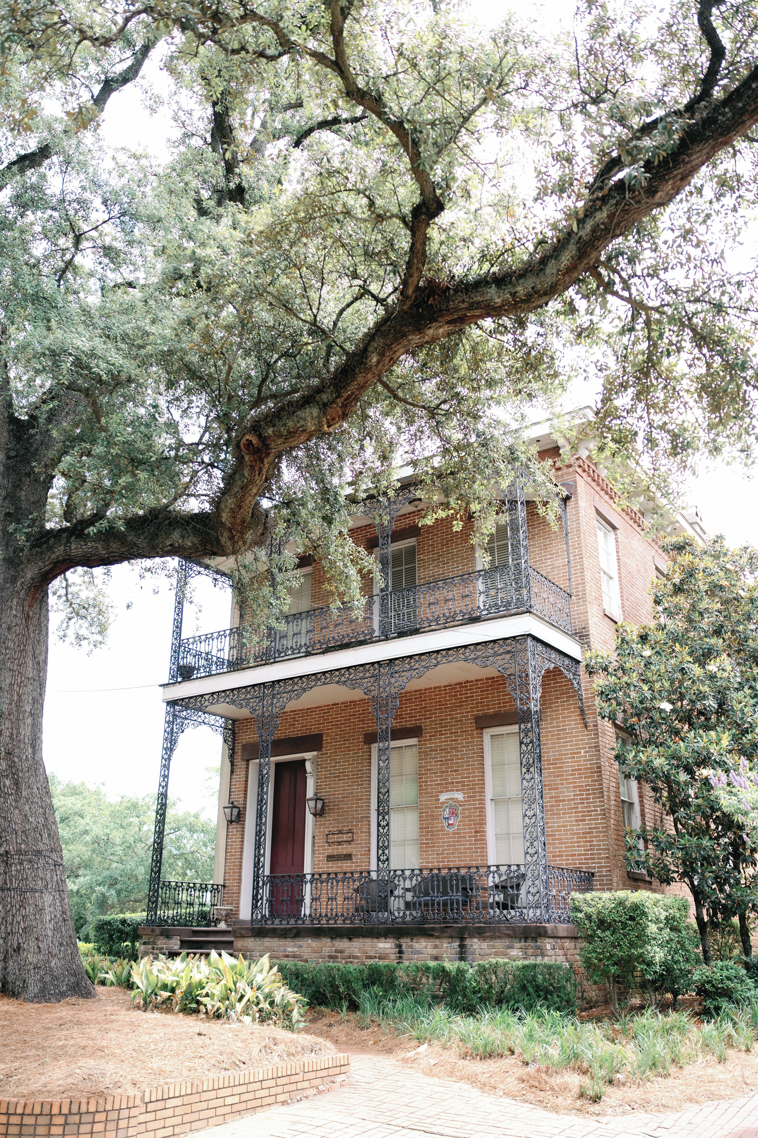 A two-story brick building with a balcony, surrounded by mature trees and a brick pathway.