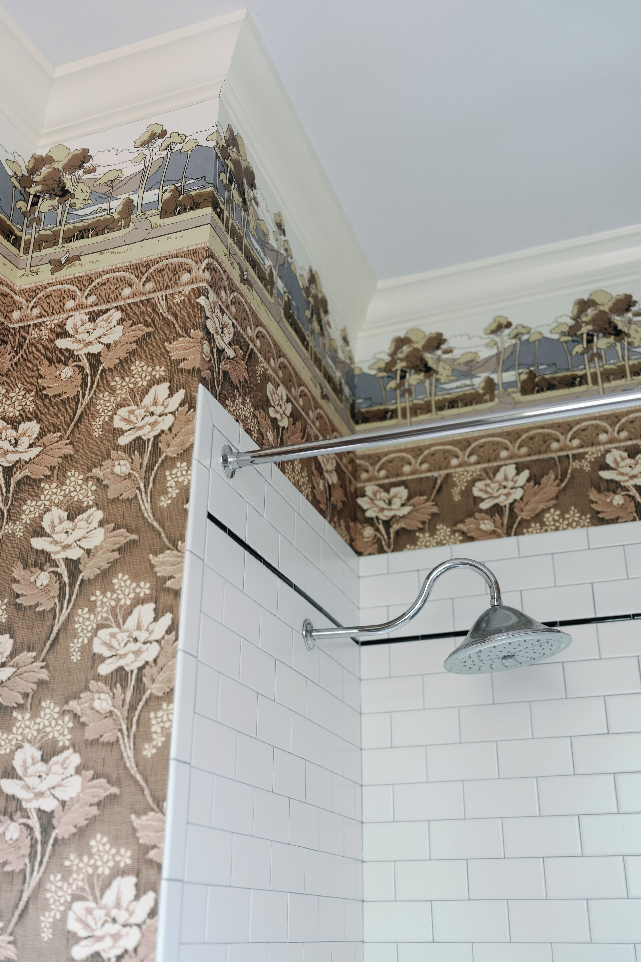 A corner of a bathroom with a shower, white tiled walls, and floral wallpaper.