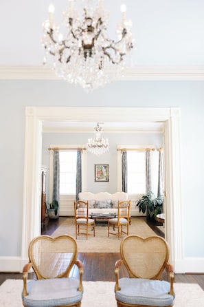 A room with a chandelier, two heart-shaped chairs, a table, and a window with curtains.