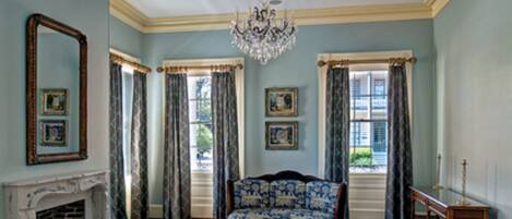 A room with a fireplace, two chairs, a blue patterned sofa, a chandelier, and a large window with curtains.