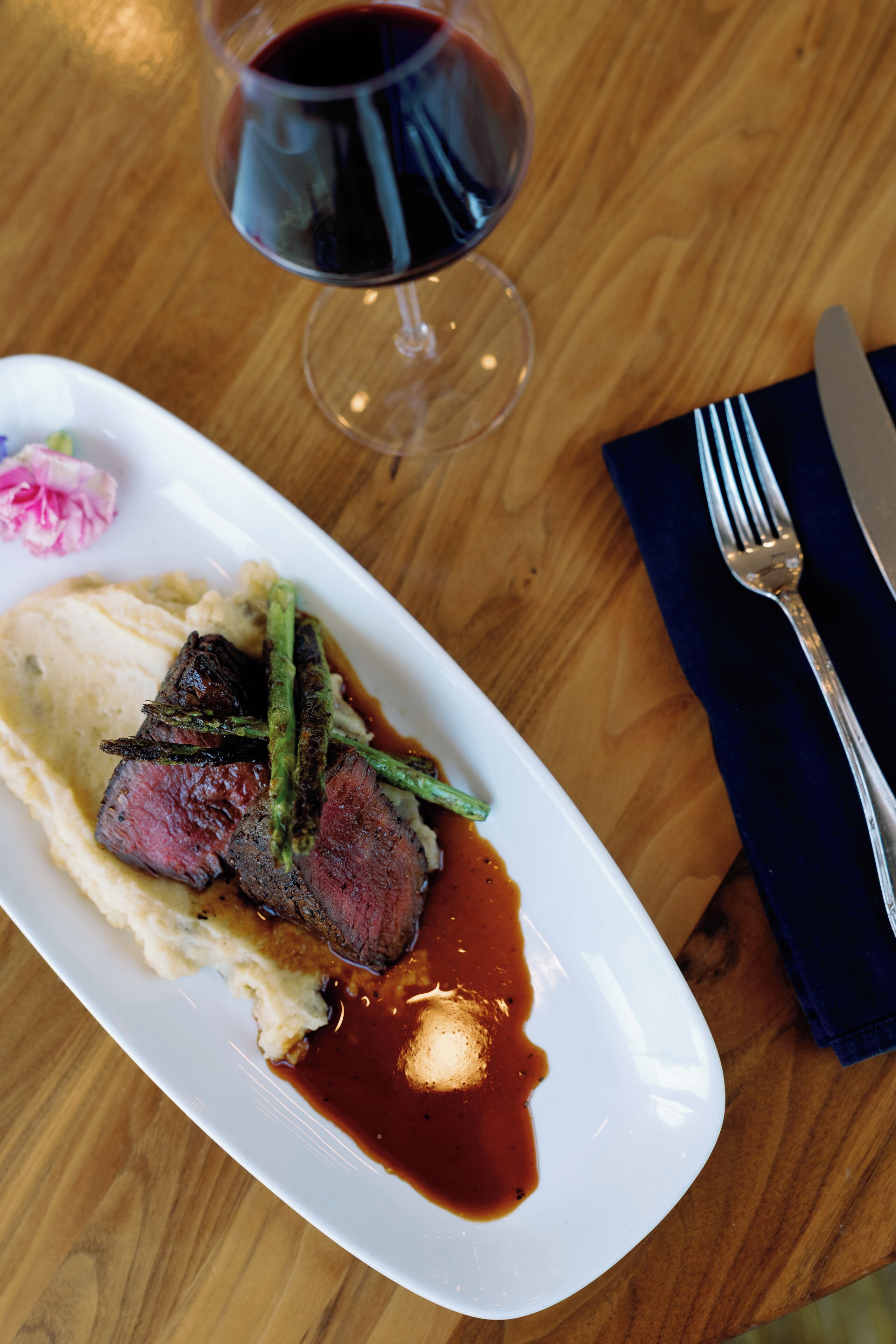 A plate of food with steak, asparagus, and sauce, a glass of red wine, and a fork and knife on a wooden table.