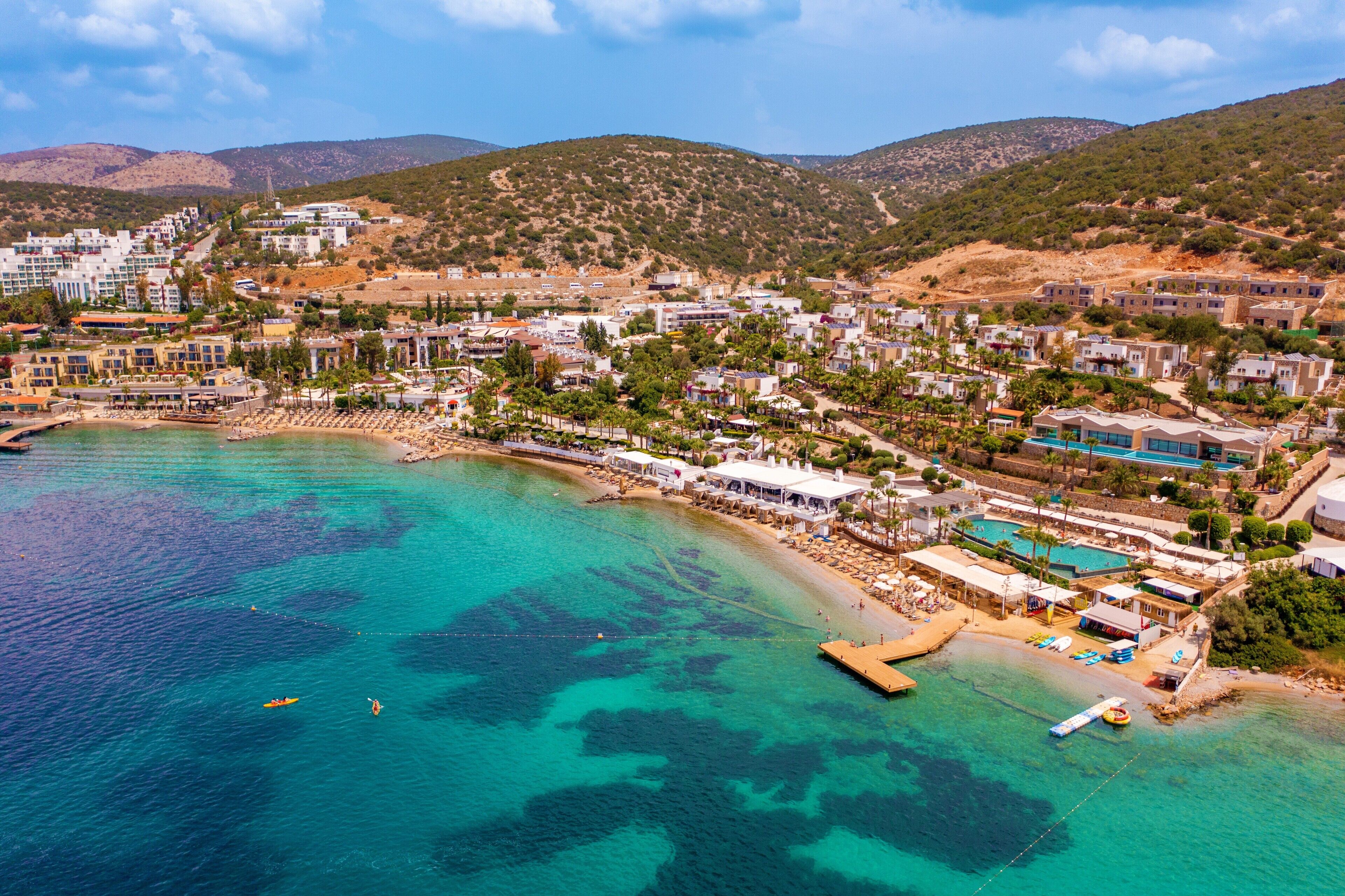 An aerial view of a coastal town with a beach, clear turquoise waters, and buildings with balconies overlooking the sea.