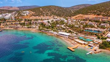 An aerial view of a coastal town with a beach, clear turquoise waters, and buildings with balconies overlooking the sea.