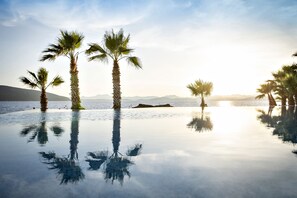 Una piscina infinita tranquila con palmeras reflejándose en el agua, rodeada de montañas y un cielo despejado.