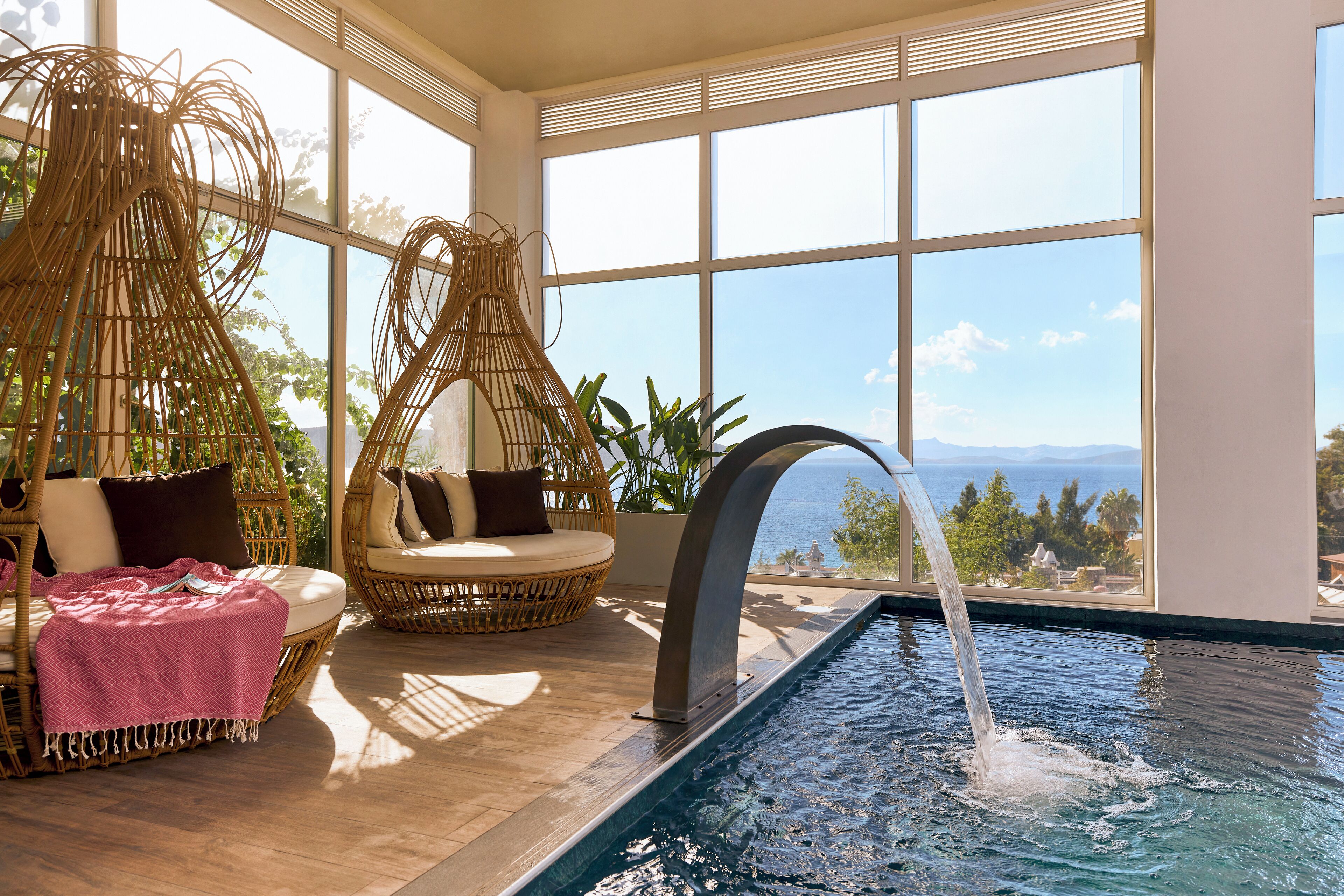 A room with a view of the ocean, a pool with a water feature, wicker furniture, and a large window.