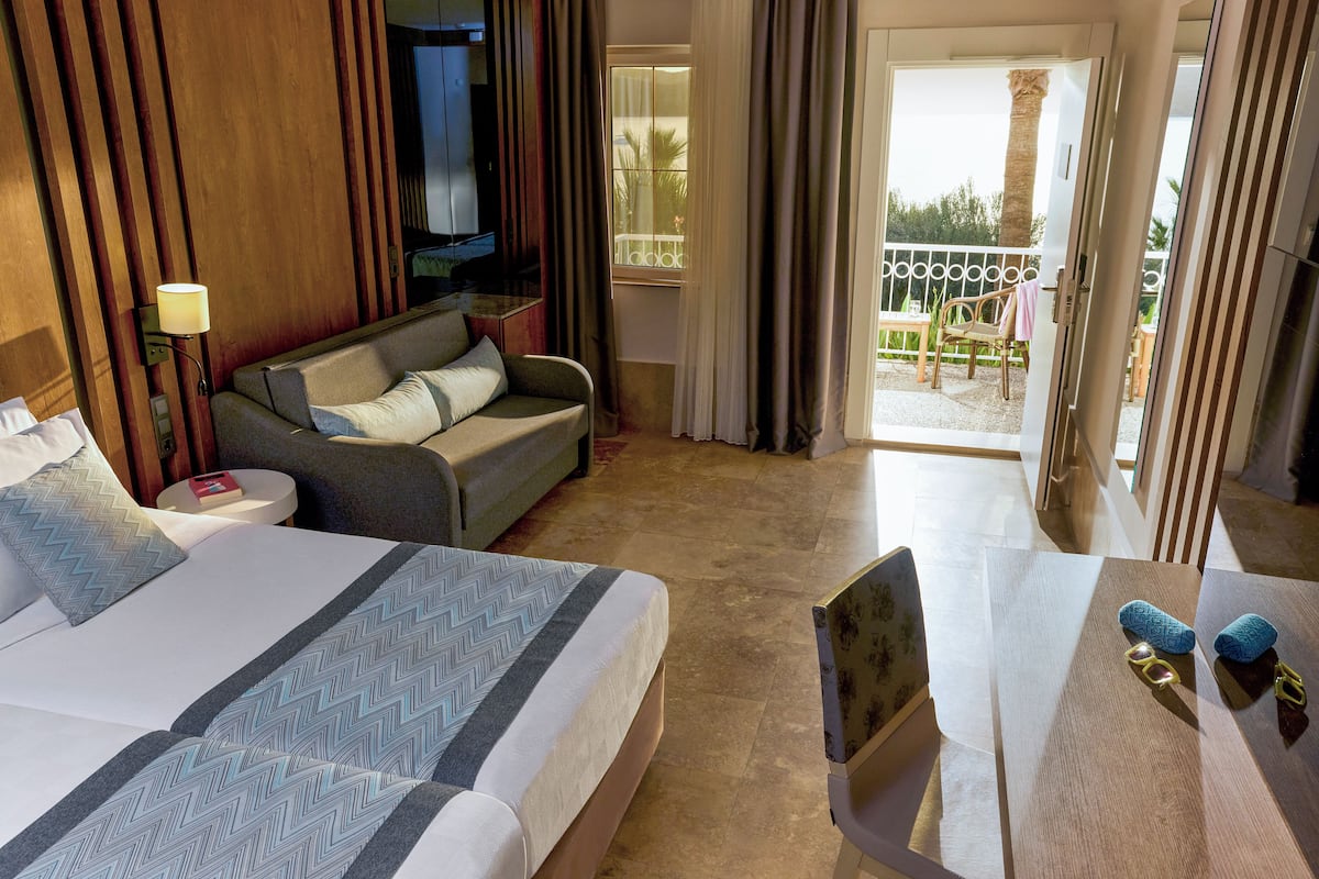 Une chambre d’hôtel avec un grand lit, un canapé, une petite table et un balcon donnant sur des palmiers.