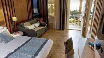 A hotel room with a large bed, a sofa, a small table, and a balcony with a view of palm trees.