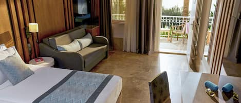 A hotel room with a large bed, a sofa, a small table, and a balcony with a view of palm trees.