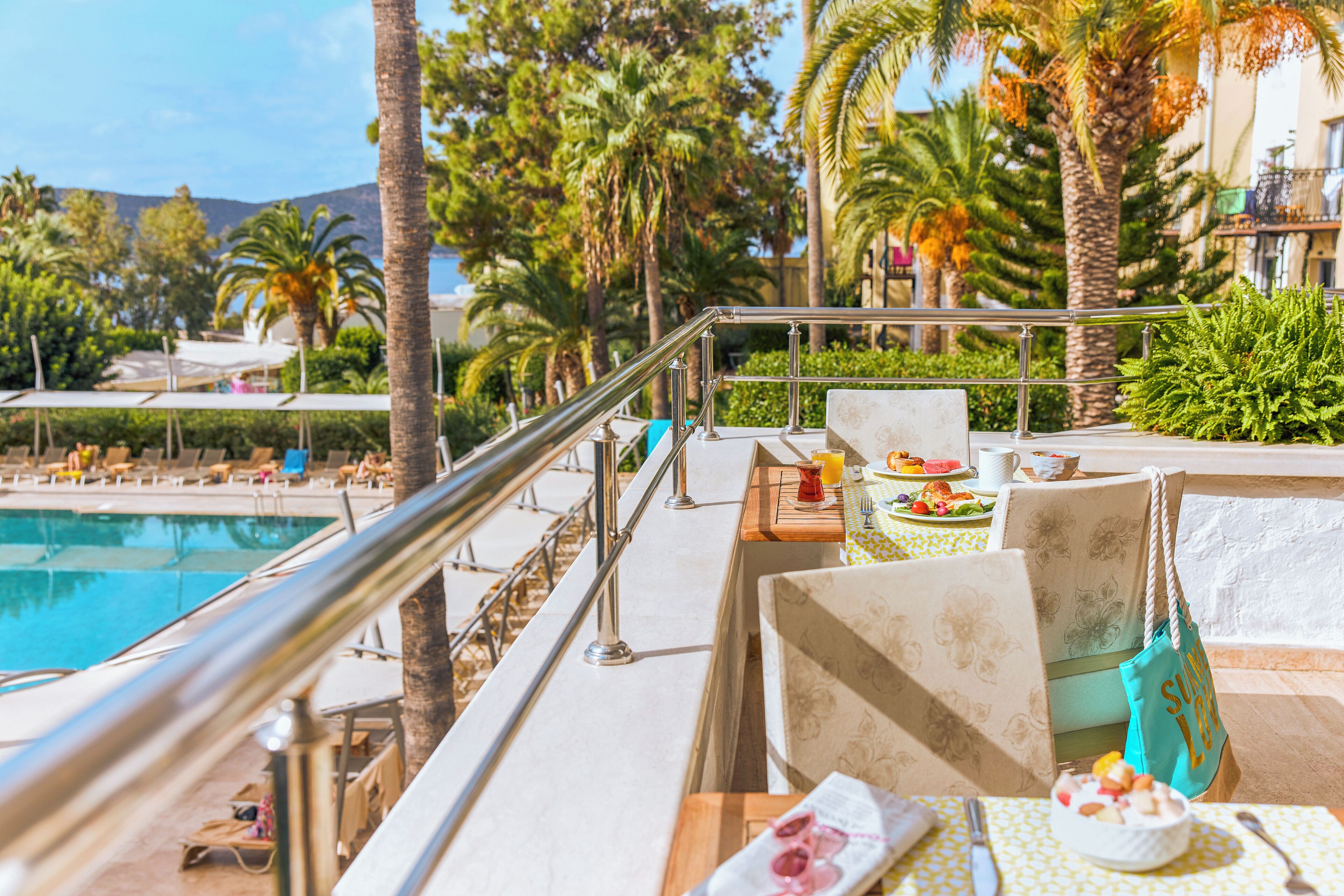 A poolside area with lounge chairs, a dining table with a meal, and a view of the pool and palm trees.