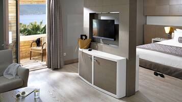 A modern hotel room with a bed, a TV mounted on the wall, a desk with a chair, and a view of the sea through a sliding door.