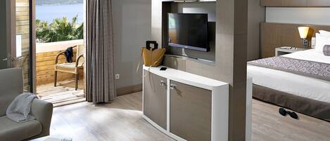 A modern hotel room with a bed, a TV mounted on the wall, a desk with a chair, and a view of the sea through a sliding door.