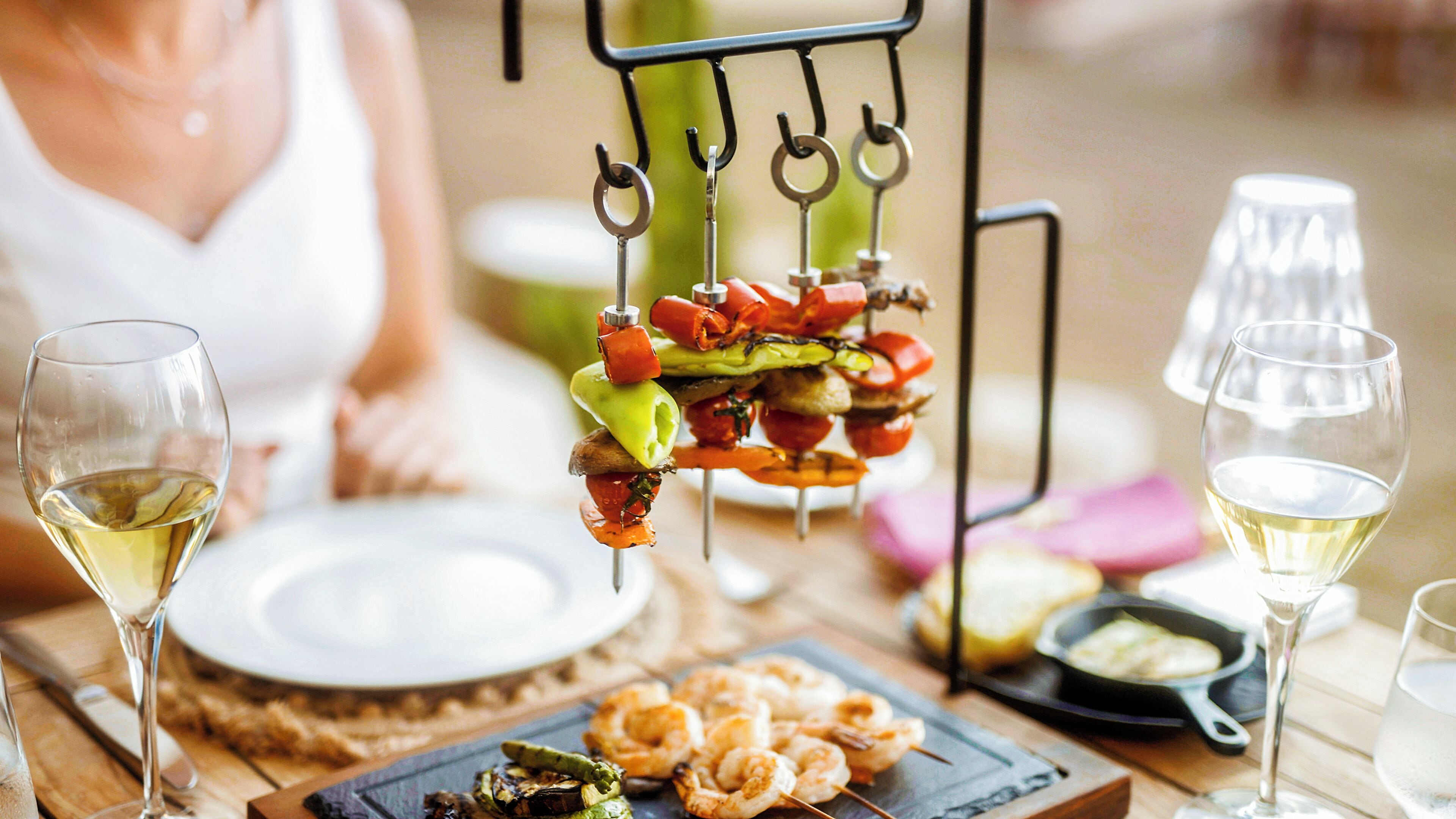 Una mesa de comedor con brochetas de verduras y camarones, un plato y copas de vino blanco.