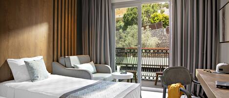 A modern hotel room with a large bed, a sofa, a small table, and a balcony with a view of greenery and a stone wall.