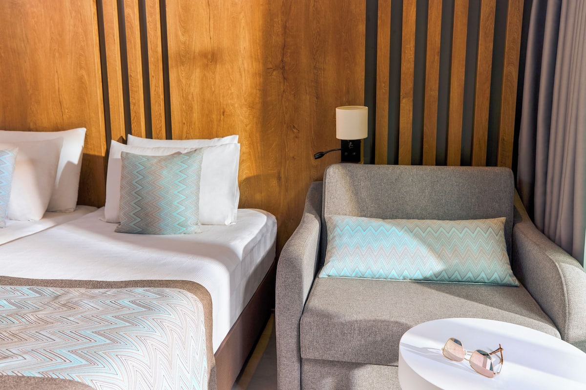 Une chambre d’hôtel avec une tête de lit en bois, deux lits avec une literie à motifs, un fauteuil gris assorti d’un coussin, et une table de chevet avec une lampe et des lunettes de soleil.