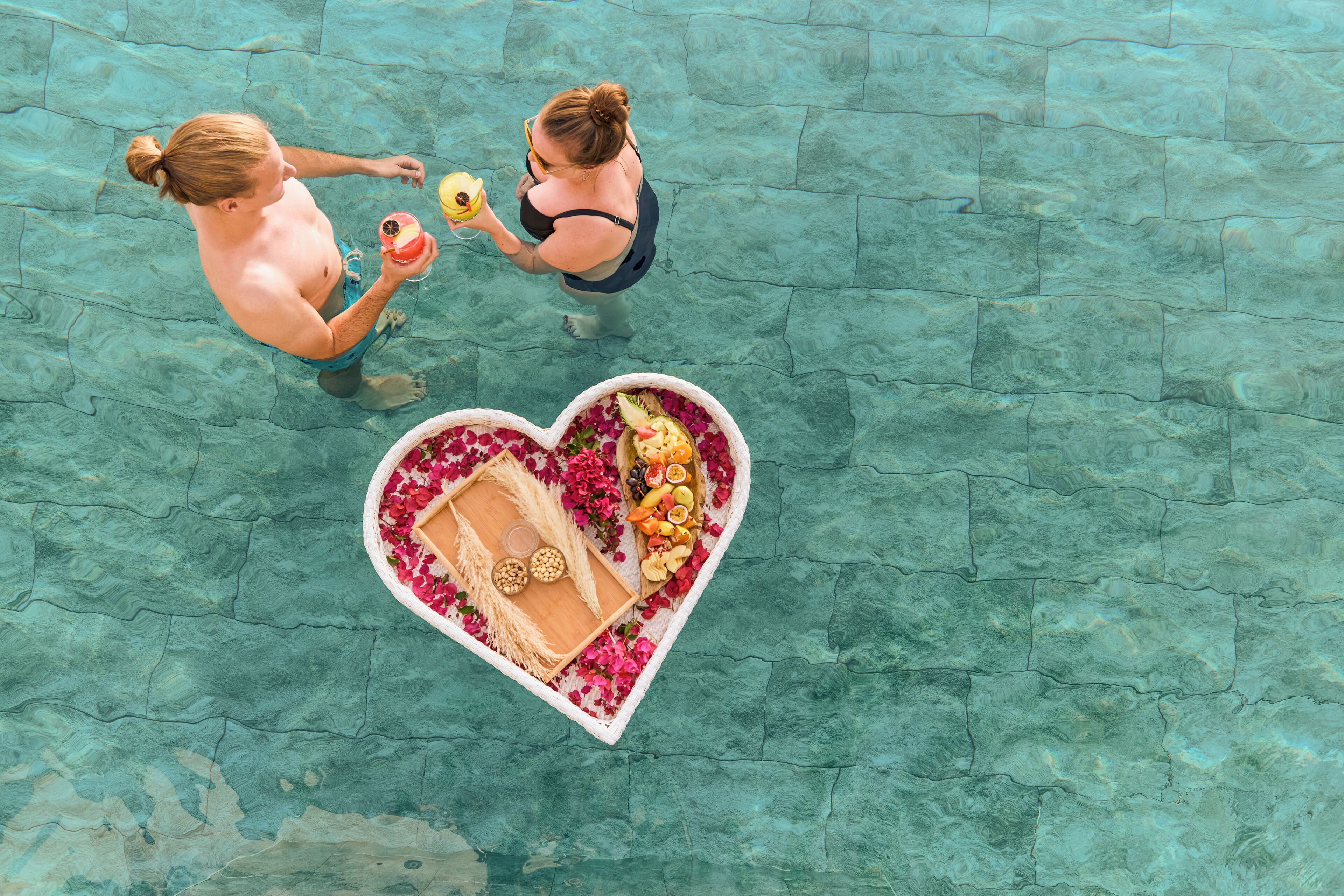 An aerial view of a swimming pool with two people conversing. A heart-shaped arrangement of flowers and fruits is placed in the pool.