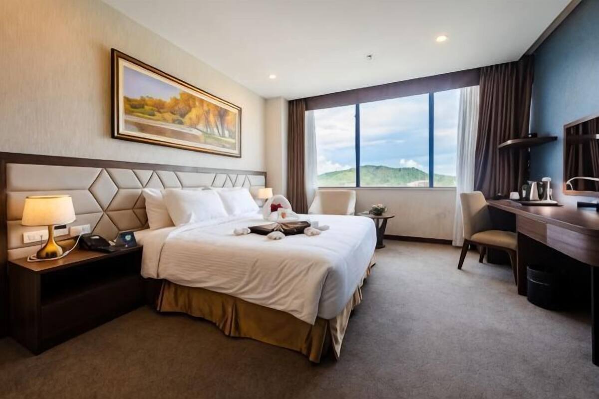 A hotel room with a bed, bedside tables, a desk, a chair, and a large window.