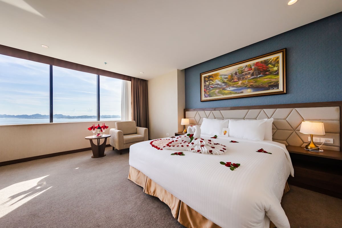 A hotel room with a large bed, a painting on the wall, a view of the sea, and a small table with a chair.
