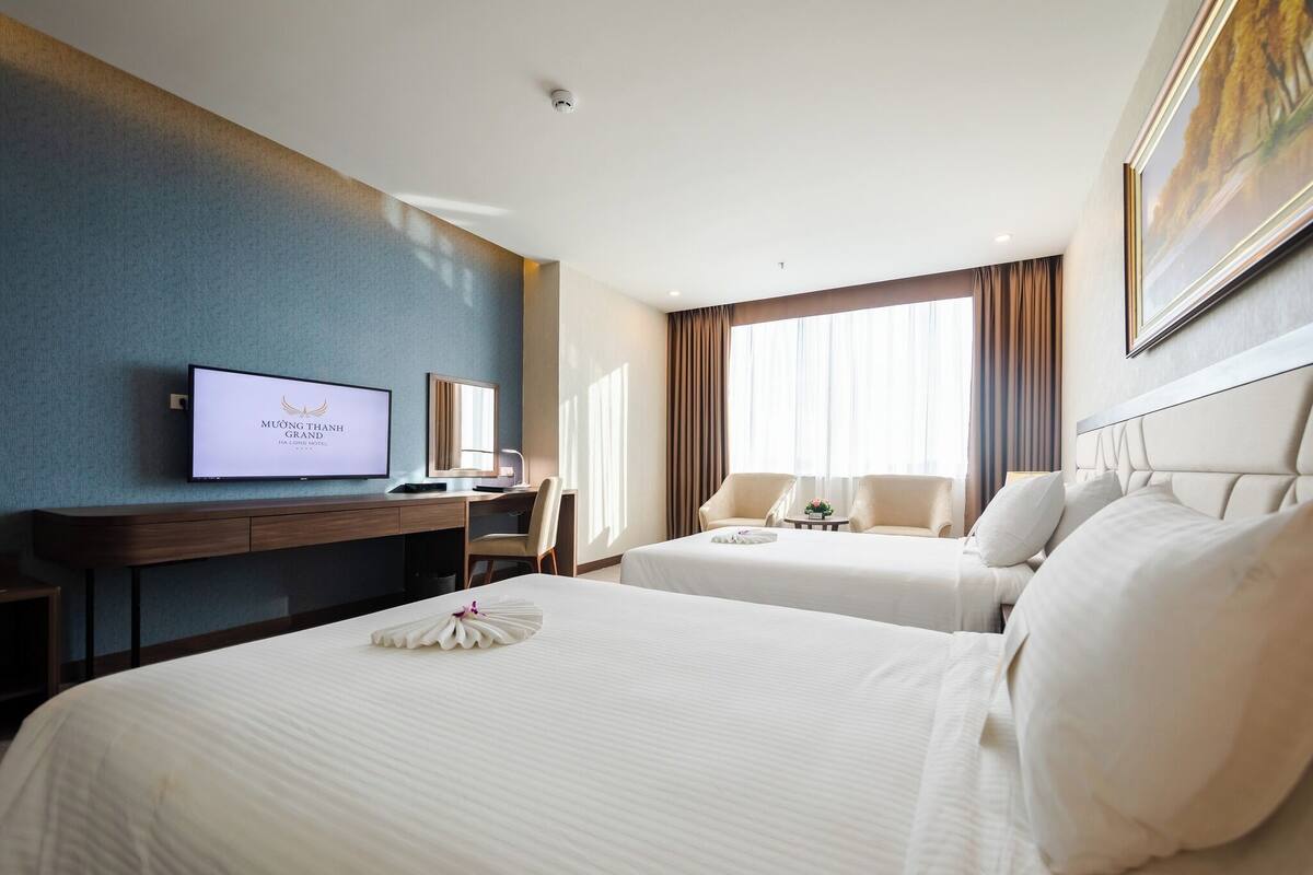 A hotel room with two beds, a desk, a television, and a large window with curtains.
