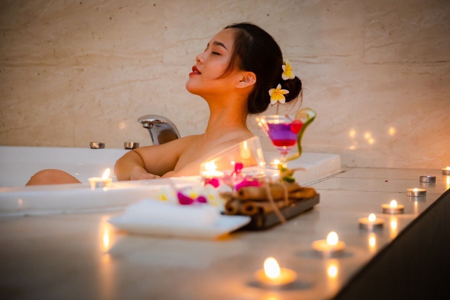 A person relaxing in a bathtub with candles and a drink on the side.