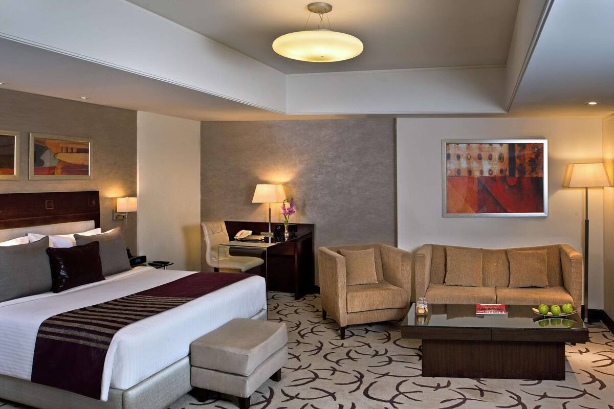 Habitación de hotel con una cama grande, un sofá, una mesa de centro, un escritorio con lámpara y un televisor.